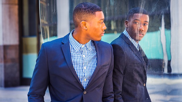 A photo of a Black man in a dark blue suit and blue and white checkered button up underneath looking at reflection of himself on a building. (Photo credit: Getty Images)