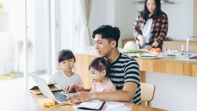 An adult works at a table with two young children watching, while another person in the household prepares food in a different part of the room. (Getty Images)