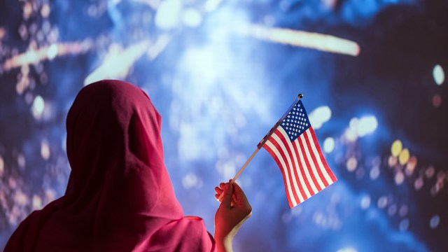 A Muslim woman in a headscarf holding an American flag during fireworks at night.