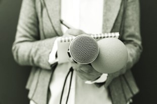 Photo showing Reporter at news or press conference, writing notes, holding microphone. Media event.